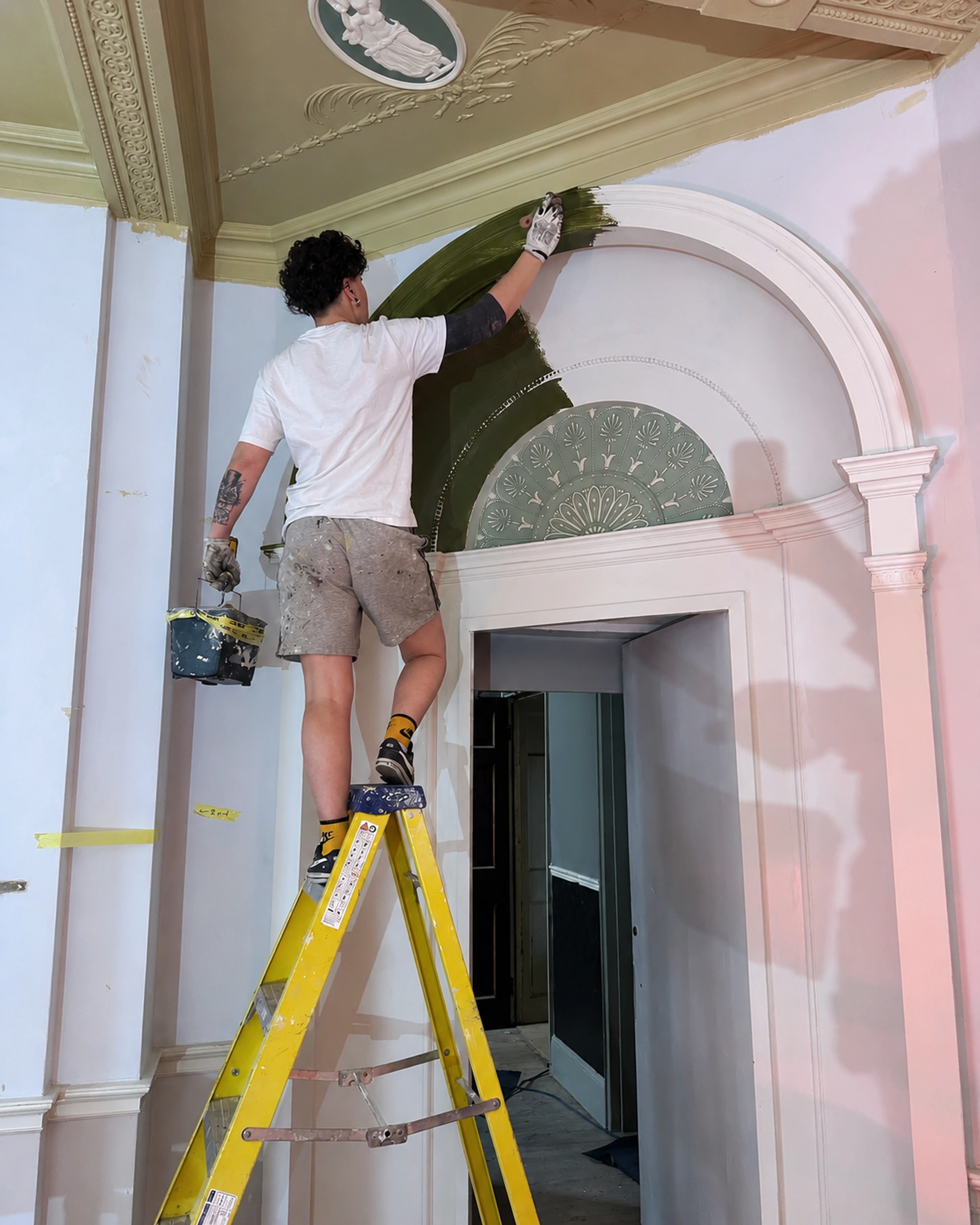 Decorator painting an ornate architectural arch olive green beneath a decorative plaster ceiling